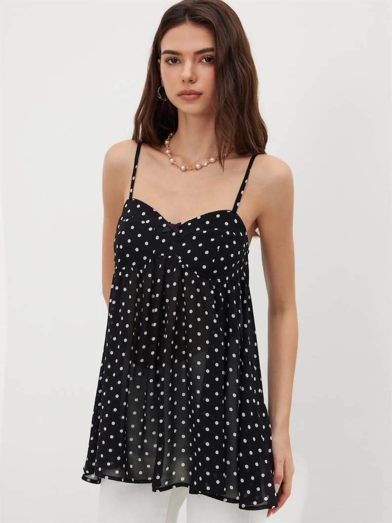 K&ouml;nnte beinhalten: Ein schwarzes Camisole-Top mit wei&szlig;en Polka-Dots. Das Top hat d&uuml;nne Spaghettitr&auml;ger und einen Sweetheart-Ausschnitt. Der Stoff wirkt leicht und flie&szlig;end. Ein stilvolles Kleidungsst&uuml;ck.