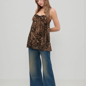 K&ouml;nnte beinhalten: Ein Outfit f&uuml;r Damen mit einem Oberteil mit Leopardenmuster und einer Jeans mit weitem Bein in Blau. Das Oberteil hat einen Sweetheart-Ausschnitt und d&uuml;nne Tr&auml;ger. Die Jeans hat eine verwaschene Optik und eine ausgestellte Silhouette. Das Outfit ist l&auml;ssig und modisch.