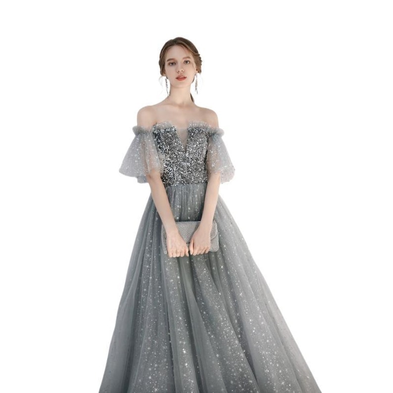 May include: A floor-length grey gown featuring an off-the-shoulder neckline with sheer tulle sleeves. The bodice is covered in silver sequins, while the skirt is made of layered tulle fabric adorned with small, shimmering silver star-shaped accents.