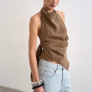 May include: A brown halter top with a draped neckline and a side tie closure, paired with light blue denim jeans. The top features a soft, flowing fabric that wraps around the torso, while the jeans have a classic five-pocket design and a light wash.