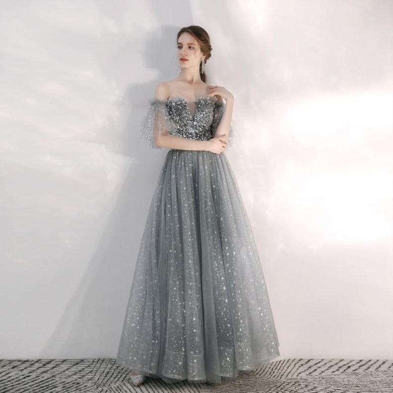 May include: A floor-length grey gown featuring an off-the-shoulder neckline with short tulle sleeves. The bodice is embellished with silver sequins, while the full skirt is crafted from layers of grey tulle adorned with a subtle star pattern.