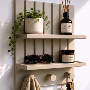 May include: A beige wall-mounted shelf unit with two horizontal shelves and two cylindrical hooks. The top shelf holds a potted trailing plant and a Meraki diffuser. The bottom shelf holds tortoiseshell sunglasses and a Black Oak candle.