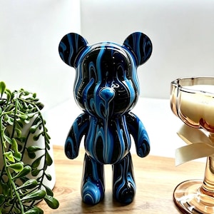 May include: A glossy bear-shaped figurine featuring a fluid, marbled pattern of deep black, royal blue, and bright white swirls. The smooth, reflective surface covers the entire body, including the rounded ears, arms, and legs, creating a continuous, abstract design that mimics liquid paint.