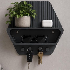 Könnte beinhalten: Ein dunkelgrauer Wand-Organizer mit gerippter Rückwand, einer oberen Ablage mit einer grünen Topfpflanze und einem weißen Kopfhörer-Etui, einem mittleren Fach mit einer schwarzen Sonnenbrille sowie vier Haken für zwei Schlüsselbunde.