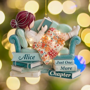 May include: A decorative hanging ornament featuring a person with dark red hair reading a book while sitting on a teal armchair. The person is covered by a floral patterned blanket. The base includes stacked books with the text Alice and Just One More Chapter.