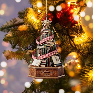 May include: A Christmas tree ornament shaped like a stack of books arranged in a conical form, wrapped in a red and white checkered ribbon. The base is a wooden hexagonal pedestal with the engraved text William 2026 and a gold star topper.