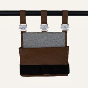 May include: A brown storage pouch with a grey fabric insert, featuring three brown straps attached to silver-toned metal buckles. A wide black horizontal band is positioned across the lower section of the pouch, which is suspended from a black rod.