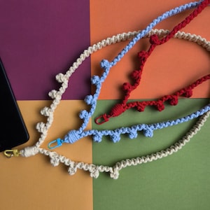 May include: Close-up of several macrame phone lanyards in cream, light blue, and red. Each lanyard has a decorative edge and a clasp for attaching to a phone. The background is a colourful patchwork of purple, orange, green, and yellow.