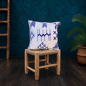 May include: A square throw pillow with a white base and blue embroidered geometric patterns, including arrows, diamonds, and zig-zag lines, resting on a small wooden stool with a woven seat and four sturdy legs.
