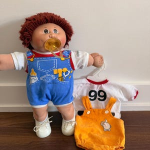 May include: A Cabbage Patch Kids doll with red hair, a dummy, and blue dungarees. The doll is wearing white shoes and a white t-shirt. Additional outfits include an orange corduroy romper and a white t-shirt with the number 99.