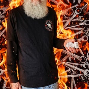 Pu&ograve; includere: Un uomo con una maglietta nera a maniche lunghe con un logo e che tiene prodotti per la cura della barba. Lo sfondo presenta un design infuocato con chiavi. I prodotti includono una latta e una bottiglia con etichette.