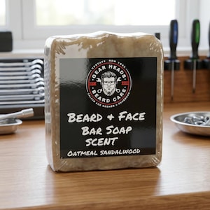May include: A rectangular bar of oatmeal sandalwood scented soap, packaged in clear plastic. The label features a logo with a man wearing glasses and the text "Gear Heads Beard Care". The soap is on a wooden surface.