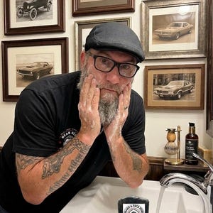 Pu&ograve; includere: Un uomo con una camicia e un berretto neri si lava il viso con una saponetta nera. Il sapone ha un'etichetta circolare con testo e grafica. Foto incorniciate di auto d'epoca sono appese al muro dietro di lui.
