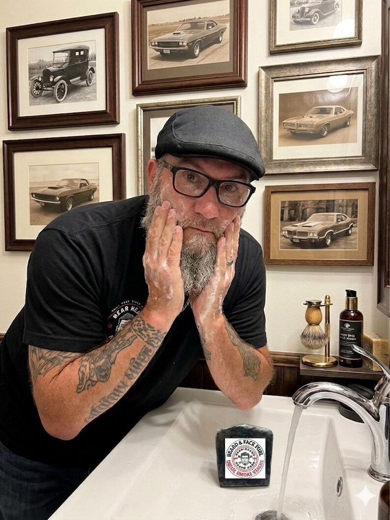Pu&ograve; includere: Un uomo con una camicia e un berretto neri si lava il viso con una saponetta nera. Il sapone ha un'etichetta circolare con testo e grafica. Foto incorniciate di auto d'epoca sono appese al muro dietro di lui.