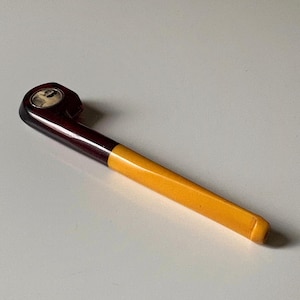 May include: A vintage tobacco pipe. The bowl and stem are dark brown, leading to a yellow-orange mouthpiece. The bowl has a small, circular inset with a black and white image. The pipe is set against a white background.