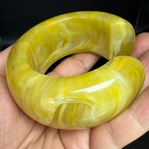 May include: A thick, open-ended cuff bracelet featuring a marbled pattern of bright yellow and creamy white swirls. The accessory has a smooth, polished finish with a rounded, chunky profile and a wide gap for sliding onto the wrist.