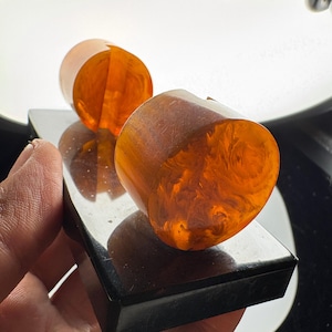 May include: Two cylindrical, translucent amber-colored resin pieces with swirling patterns are mounted onto a rectangular, polished black base. The resin objects feature a vertical slit cut into their centers, creating a distinct, smooth, and glossy appearance.