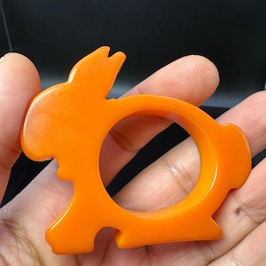 Puede incluir: Servilletero con forma de conejo de color naranja brillante. El anillo está hecho de un material sólido y liso, con una abertura circular en el centro. El conejo tiene orejas largas y una cola pequeña. Un artículo divertido y decorativo para la mesa.