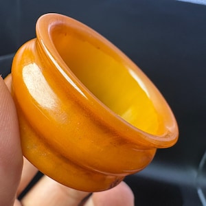 Puede incluir: Primer plano de un anillo translúcido de color ámbar. El anillo tiene un exterior liso y pulido con un interior amarillo dorado cálido. El diseño del anillo presenta una forma redondeada con un ligero labio en el borde exterior.