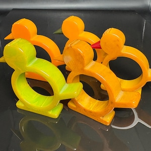 Op de afbeelding: Een set van vier vogelvormige servetringen in tinten van feloranje, geelgroen en gemarmerd amber. Elke ring heeft een ronde opening, een puntige snavel en een platte voet, waarbij één vogel een opvallend rood accent op de snavel heeft.