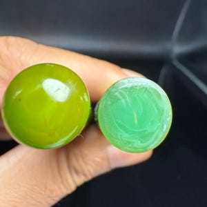 Puede incluir: Dos objetos cilíndricos con extremos planos y circulares. El objeto de la izquierda presenta una superficie verde oliva lisa y translúcida. El objeto de la derecha muestra una superficie verde menta opaca más clara con sutiles patrones de mármol blanco.