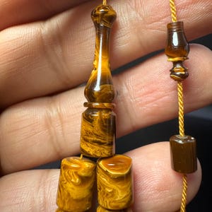 Könnte beinhalten: Nahaufnahme eines Gebetsketten-Sets mit einem marmorierten bernsteinfarbenen und braunen Design. Die Perlen sind auf einer goldfarbenen Schnur aufgefädelt. Das Set enthält ein langes, verziertes Mittelstück und mehrere zylindrische Perlen.