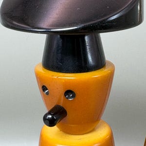 May include: A vintage wooden toy figure with a black hat, nose, and details. The figure has an orange body and yellow base. The eyes are represented by small, silver-coloured rivets. The toy is a unique, handcrafted item.