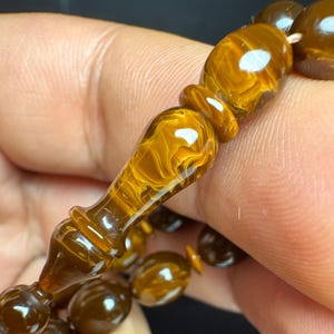 May include: A prayer bead strand featuring smooth, polished amber-colored beads with a translucent, swirling marbled pattern. The central bead is elongated and shaped like a spindle, while the surrounding beads are rounded and oval-shaped.