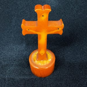 May include: An orange-colored wooden cross with a detailed design, mounted on a round base. The cross has a classic shape with flared ends and is set against a dark background. The base is also orange, with a lighter-colored ring.