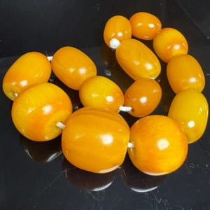 Può includere: Una collana composta da perline lisce, levigate e a forma di barilotto in varie tonalità di arancione vibrante e giallo dorato. Le perline sono infilate su un cordino bianco e presentano una finitura lucida, semitraslucida e marmorizzata.