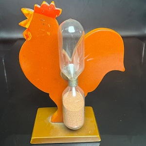 Könnte beinhalten: Ein dekorativer, orangefarbener Ständer in Hahnform mit einer klaren Sanduhr aus Glas in der Mitte. Die Sanduhr enthält feinen beigen Sand in der unteren Glaskugel. Das gesamte Objekt steht auf einem massiven, rechteckigen senfgelben Sockel.