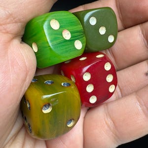 Puede incluir: Un conjunto de cuatro dados de seis caras en varios colores, incluyendo verde brillante con textura veteada, verde oliva sólido, rojo intenso y amarillo mostaza con acabado veteado. Cada dado presenta esquinas redondeadas y puntos blancos.