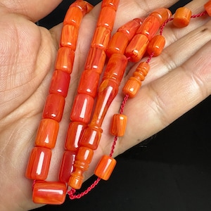 Puede incluir: Un rosario de cuentas cilíndricas de color naranja con textura marmolada, ensartadas en un cordón rojo fino. El rosario incluye una cuenta central alargada con estrías decorativas y una serie de cuentas naranjas uniformes, lisas y pulidas.