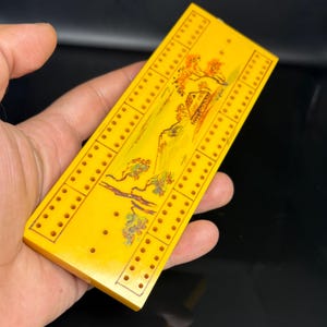 Puede incluir: Un tablero de cribbage rectangular de color amarillo con una ilustración pintada a mano de un paisaje con una casa, árboles y montañas. El tablero tiene tres pistas paralelas de agujeros perforados para anotar puntos.
