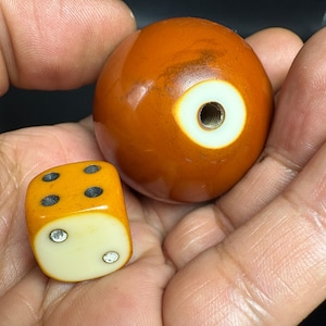 May include: A smooth, spherical, amber-colored object with a central white circular inlay featuring a threaded metal hole, positioned next to a small, cube-shaped die with a yellow body, a white face showing two silver pips, and black pips on top.