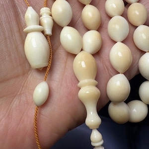May include: Ivory-coloured prayer beads with an ornate design. The beads are oval-shaped and strung together with a gold-coloured cord. The focal point features a decorative, elongated bead with a bulbous shape. The beads are smooth and have a glossy finish.