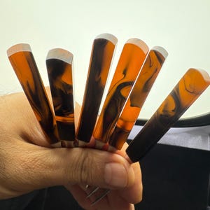 May include: Six orange and black marbled pens with white tips are held in a hand. The pens have a rectangular shape and a glossy finish. The pens are arranged in a fan shape, with the tips facing upwards.