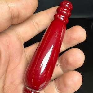 Op de afbeelding: Een glad, glanzend, dieprood cilindervormig object met een taps toelopend lichaam en een afgerond, geribbeld bovengedeelte. Het stuk heeft een gepolijste afwerking en een solide, uniforme kleur over de gehele langwerpige, licht bolvormige structuur.