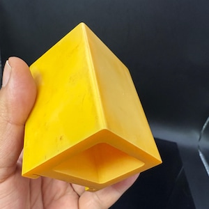 May include: A bright yellow, geometric, hollow cube with a square opening at the bottom. The cube is held in a hand against a dark background. The cube's edges are sharp and well-defined.