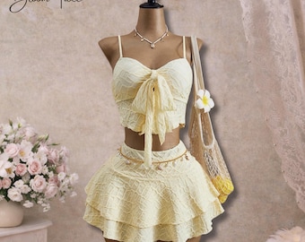 May include: A two-piece yellow outfit on a mannequin. The top is a crop top with a bow and spaghetti straps. The skirt is a mini skirt with ruffles. A necklace and a small bag with a flower are also included. The text "Bloom Tide" is in the upper left corner.