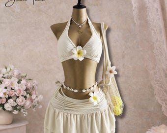 May include: Cream-colored two-piece swimsuit with a halter top and skirt, adorned with white flowers. A seashell necklace and a woven bag with a flower and yellow fruit complete the beach-ready ensemble. The text "Bloom Tide" is in the upper left corner.