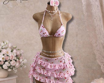 May include: A pink gingham bikini set with a cherry print, displayed on a mannequin. The set includes a halter top, a ruffled skirt, and a matching floral hair accessory. Delicate necklaces and a waist chain complete the ensemble. The text "Bloom Tide" is visible.