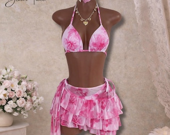 May include: Pink floral print bikini set with a matching tiered skirt. The set includes a halter top, skirt, and a floral hair accessory. A gold necklace with a heart pendant is also included. The text "Bloom Tide" is in the upper left corner.