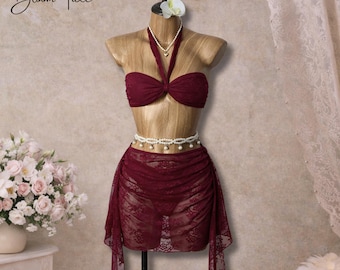 May include: A burgundy lace bikini set displayed on a mannequin. The set includes a halter top, a high-waisted skirt, and a pearl necklace. The words "Bloom Tide" are written in cursive in the upper left corner.
