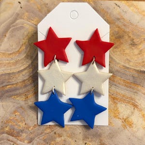 May include: A pair of dangle earrings featuring three stacked star shapes in red, white, and blue. Each earring consists of a solid red star at the top, a shimmering white star in the middle, and a solid blue star at the bottom, attached to a white card.