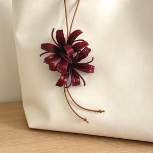 Può includere: Una borsa tote color crema con un ornamento floreale rosso scuro realizzato con sottili strisce di pelle. Il fiore è fissato a un lungo e sottile cordino di pelle marrone che pende sulla parte anteriore della borsa e termina con due piccoli nodi.