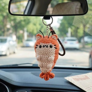 May include: A handmade orange and cream-colored crochet shrimp car charm hanging from a rearview mirror. The shrimp has black eyes, whiskers, and a brown leather strap. The charm is suspended in front of a car dashboard.