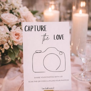 Op de afbeelding: Een witte kaart met de tekst "CAPTURE the LOVE" en een camera-illustratie. De kaart bevat ook instructies om foto's te delen via een QR-code. De kaart wordt vastgehouden door een goudkleurige basis, waarschijnlijk voor een bruiloft of evenement.