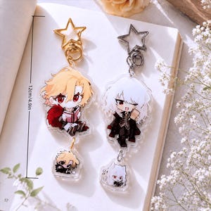 Könnte beinhalten: Zwei Acryl-Schlüsselanhänger mit Anime-Figuren und sternförmigen Verschlüssen. Der linke Anhänger hat einen goldenen Verschluss und eine blonde Figur, die ein Buch liest, der rechte einen silbernen Verschluss und eine weißhaarige Figur. Länge: 12 cm.