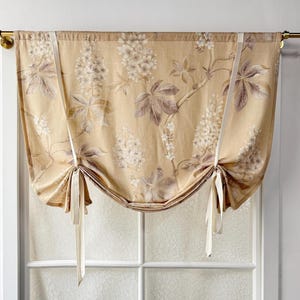 Puede incluir: Una cortina romana beige con un estampado floral de flores y hojas blancas y marrones. La cortina está atada con cintas color crema y cuelga de una barra dorada sobre una ventana.